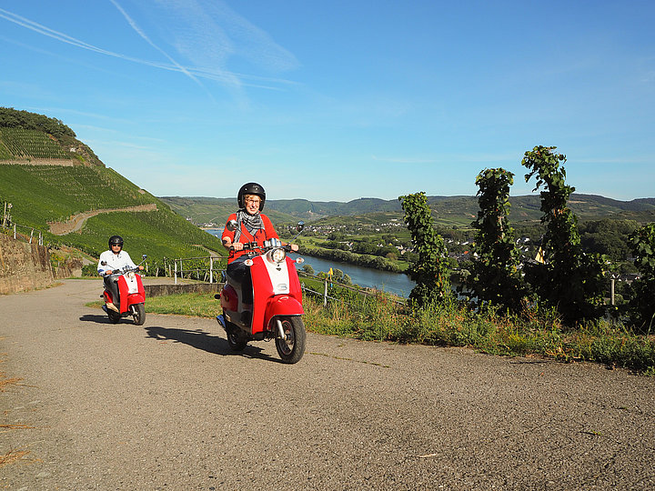 Das Bild zeigt zwei Personen, die mit roten Vespa-Rollern durch die Weinberge bei Zeltingen-Rachtig fahren.