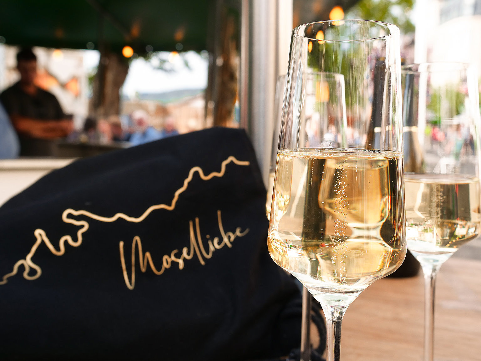 Das Bild zeigt zwei Sektgläser, die auf einem Tisch stehen. Auf dem Tisch steht weiterhin ein Aufsteller auf dem "Moselliebe" steht. Im Hintergrund ist das Weinfest der Mittelmosel, genauer die Weinstraße zu sehen.