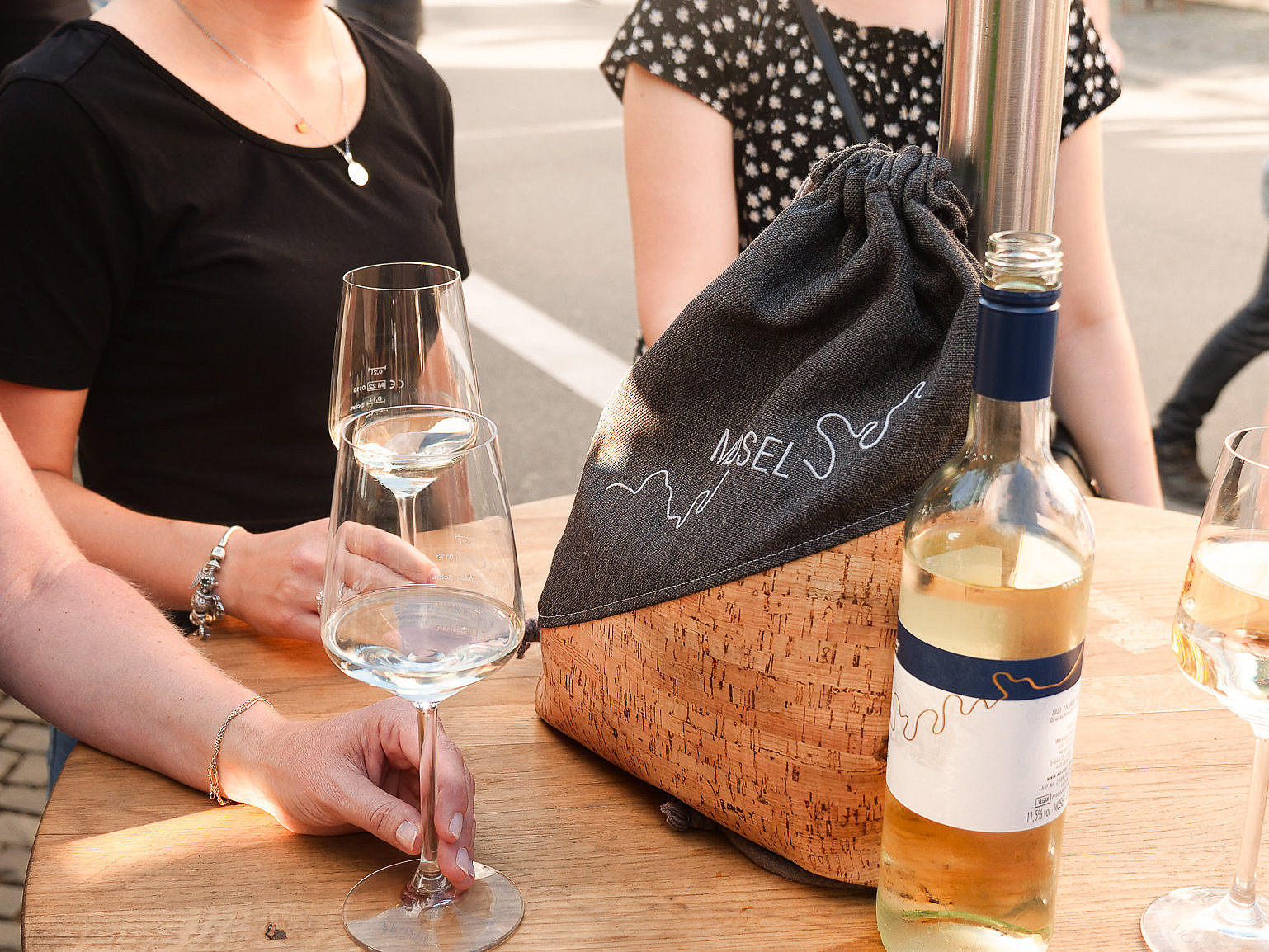 Das Foto zeigt Personen, die an einem Tisch stehen und Wein trinken. Auf dem Tisch ist eine Weinflasche und ein Turnbeutel mit der Aufschrift Moselliebe zu sehen.