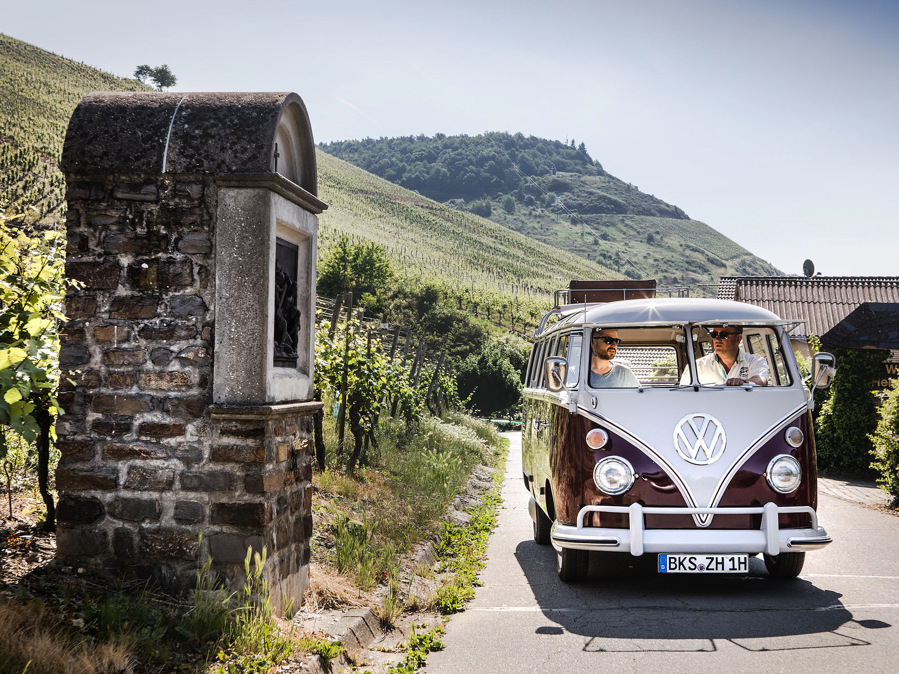 Das Bild zeigt einen VW Bulli Oldtimer, der durch die Moselweinberge fährt.