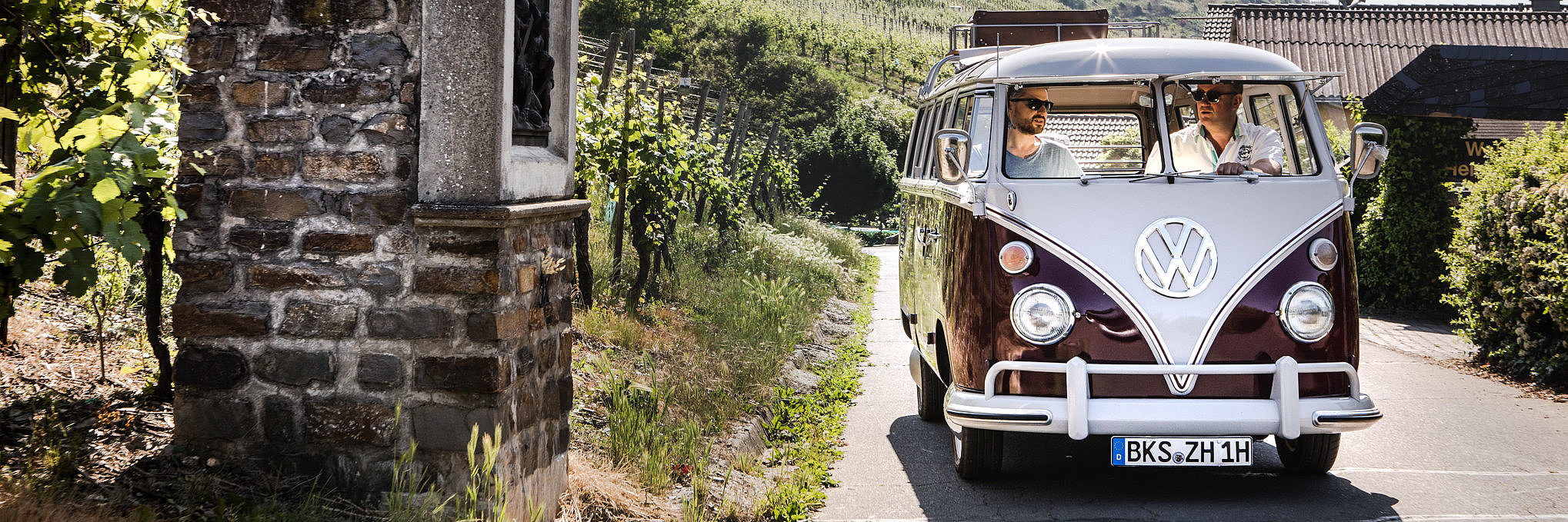 Das Bild zeigt einen VW Bulli Oldtimer, der durch die Moselweinberge fährt.
